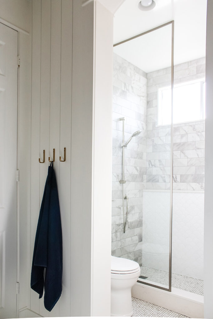 Vertical shiplap bathroom walls and Magnolia line from Target brass hooks, scallop tile shower ledge wall, marble subway tile, glass shower divider, polished nickel fixtures.