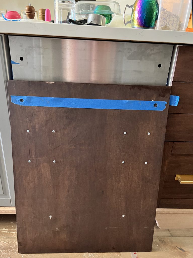 blue tape with the measurements and markings from the stainless steel panel attached to the diy faux dishwasher panel to line up the initial screws for joining the two together