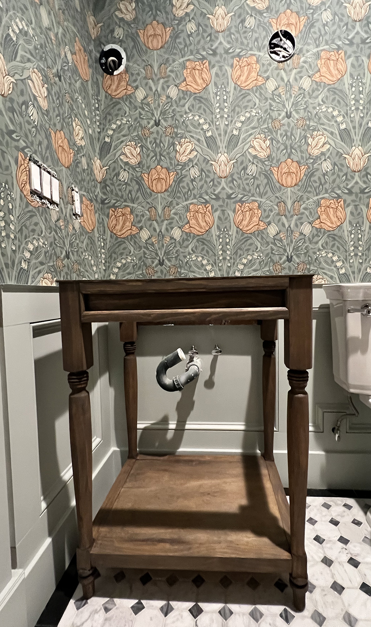medium wood stained vanity in a weathered finish placed in front of sage green floral wallpaper and a diy wainscoting panel wall